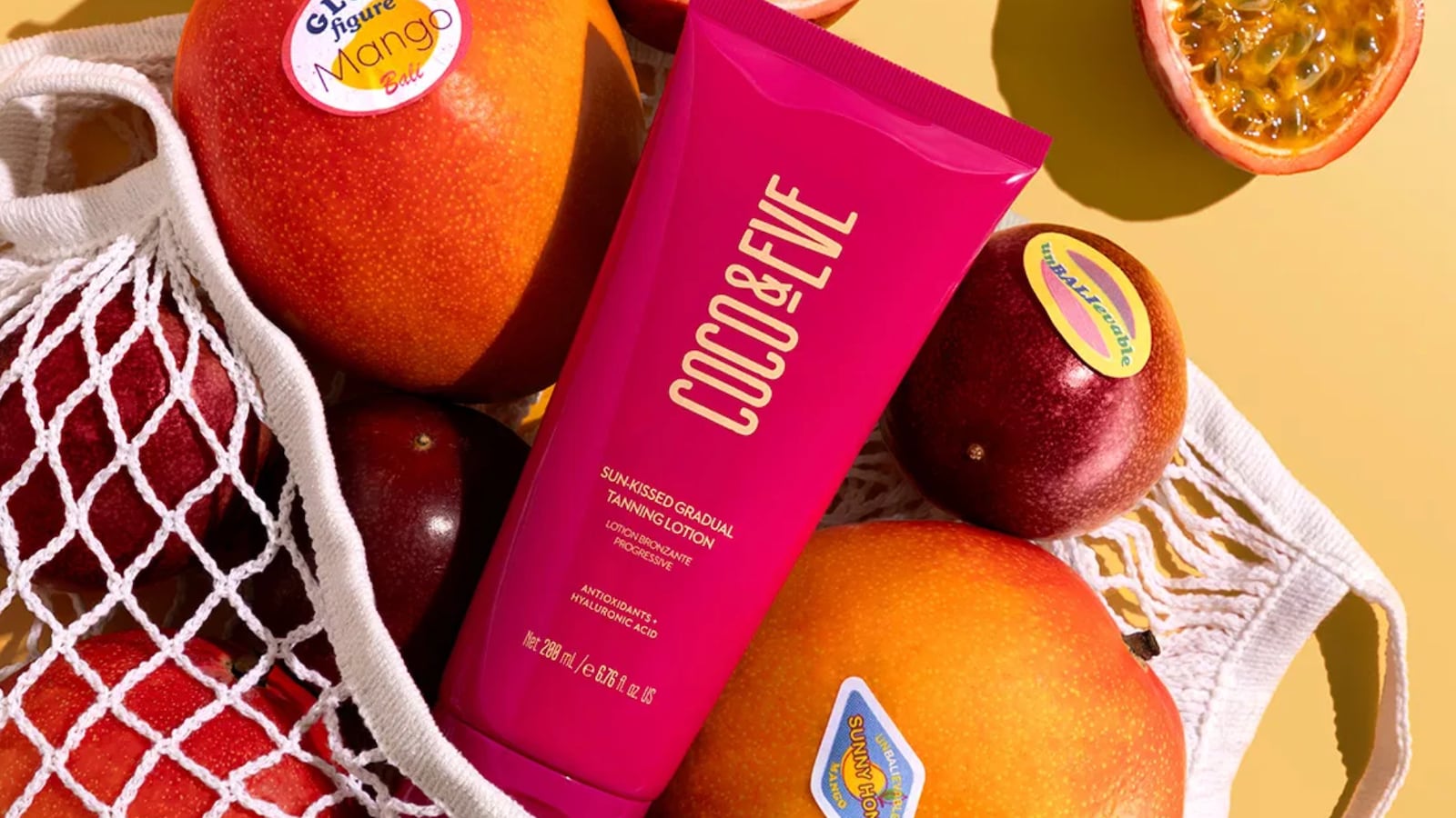 A fuchsia bottle of Coco & Eve's tanning lotion. It's in a white bag filled with mango fruits.