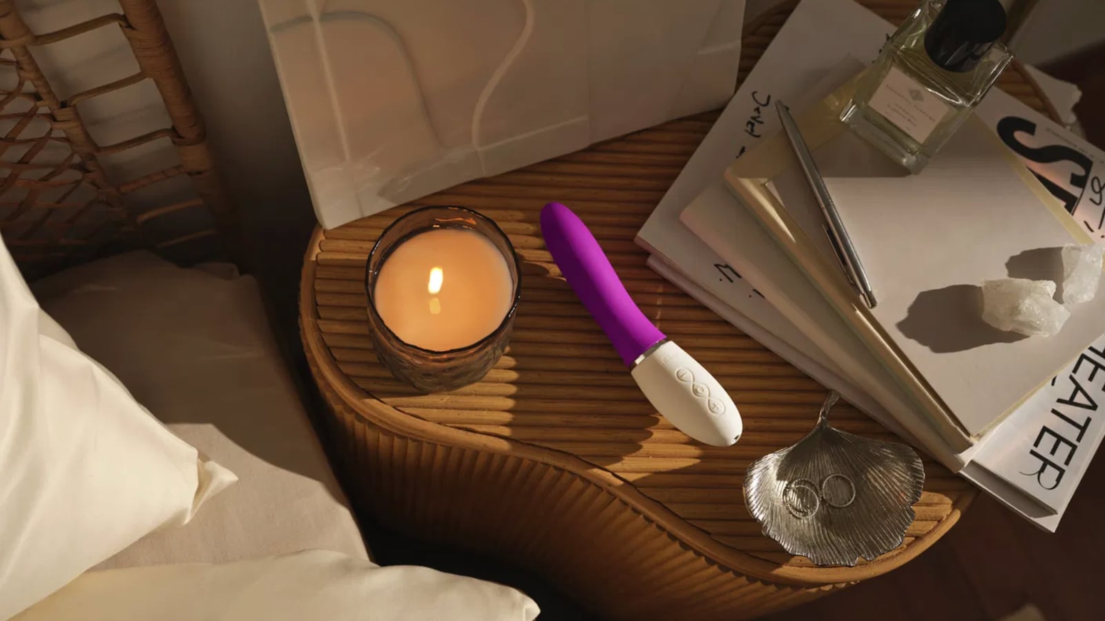 One a LELO's vibrators laying on a bedside table. The vibrator is purple and white. Next to the vibrator is a lit candle, seashell, and a stack of books.