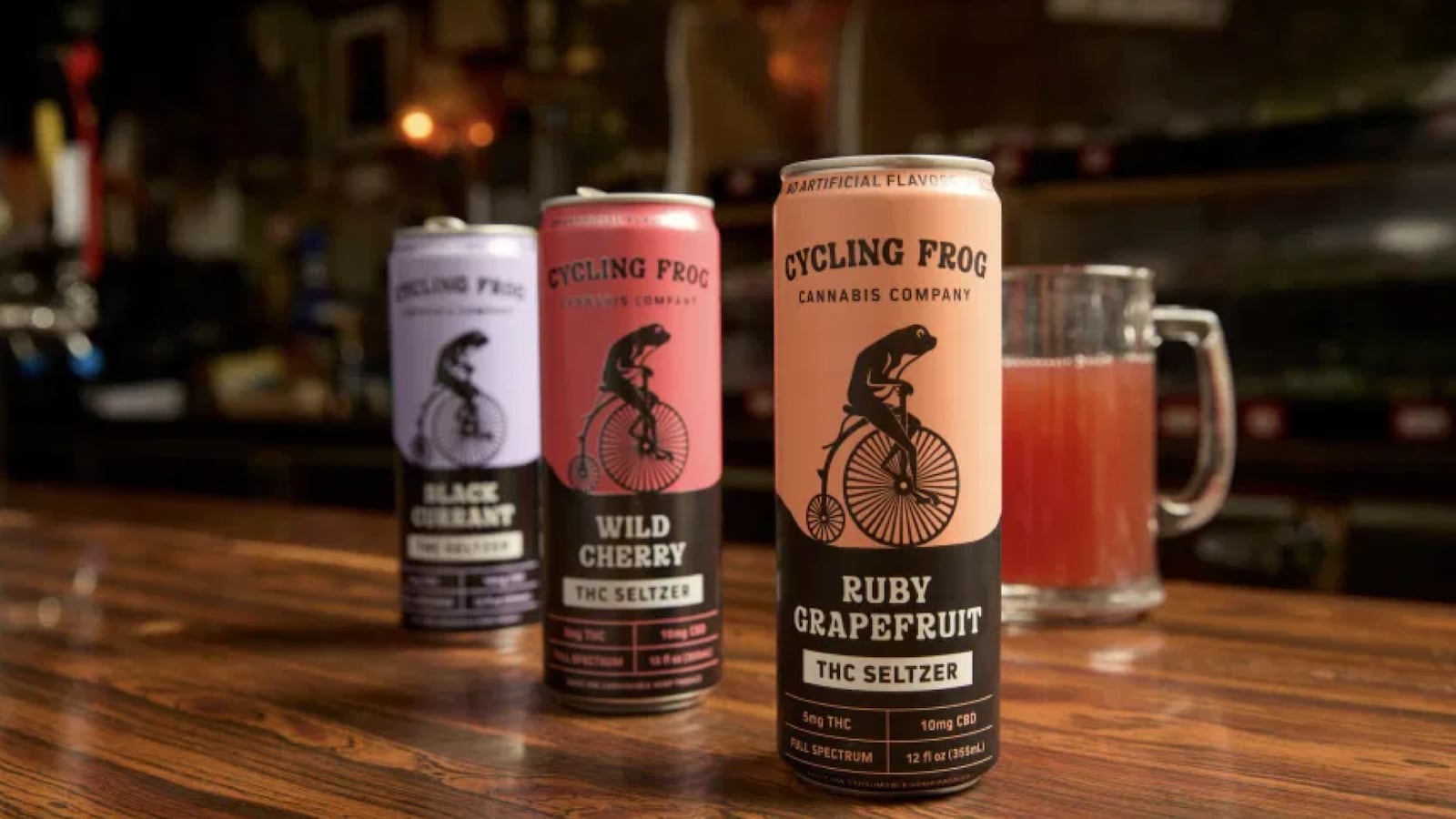 A bar top with three cans of Cycling Frog seltzer and a mug with a orange-colored drink.
