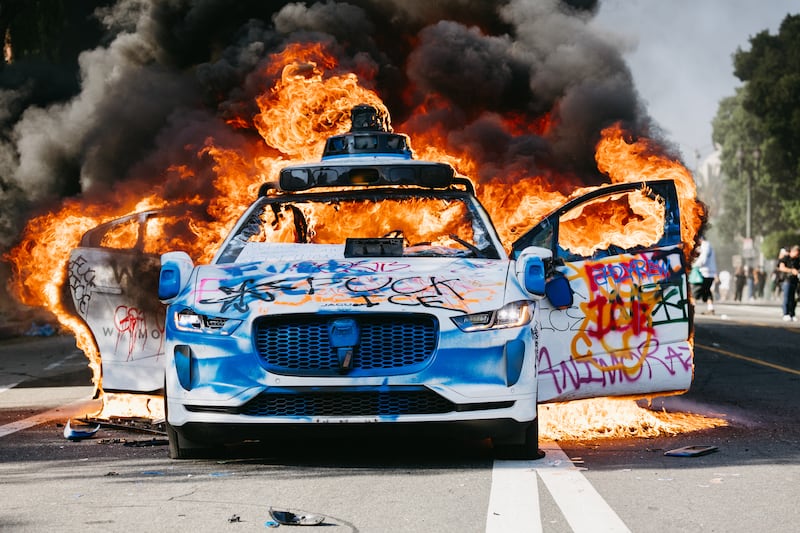 A Waymo car burns in the street as smoke billows during protests in Los Angeles, California, on June 8, 2025. Following a series of aggressive federal immigration operations in Los Angeles, tensions escalated when Immigration and Customs Enforcement (ICE) agents conducted a high-profile raid on a Home Depot location. The raid sparked widespread protests across the city, where demonstrators decried the targeting of immigrant communities and the separation of families. Clashes soon broke out between protesters and federal agents, prompting U.S. President Donald Trump to deploy 2,000 National Guard troops to the city, a move harshly criticized by California officials, including the governor, as "purposefully inflammatory." (Photo by David Pashaee / Middle East Images via AFP) (Photo by DAVID PASHAEE/Middle East Images/AFP via Getty Images)