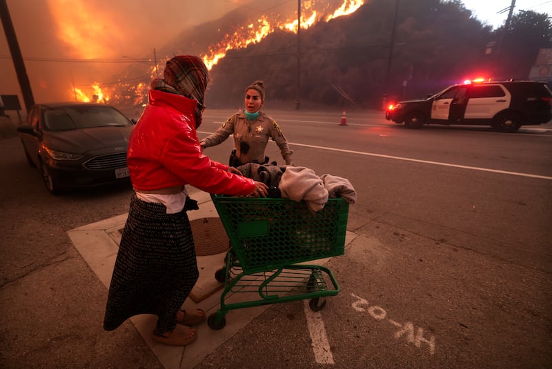 A police officer escorts a homeless woman to evacuate away from Pacific Coast Highway and Topanga Canyon Blvd as the Palisades Fire rages down the hills in Pacific Palisades, Calif. on Tuesday, Jan. 7, 2025.