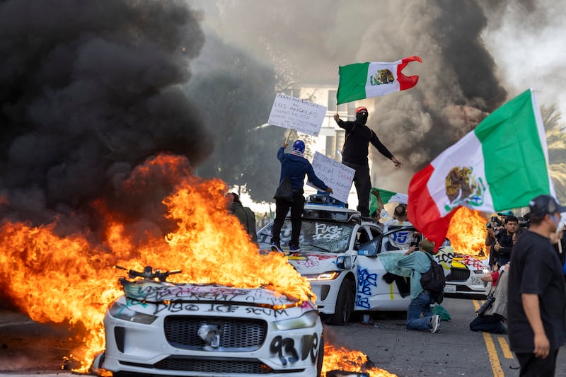 Demonstrators wave flags from atop a wrecked car, standing beside burning Waymo vehicles.