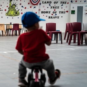 An immigrant child plays in front of patriotic phrases and symbols covering the walls in the gymnasium of an ICE facility.