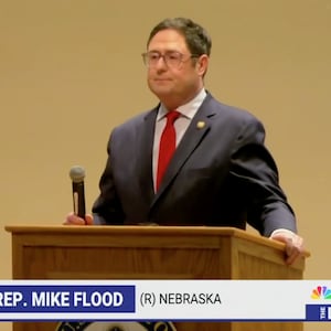 Rep. Mike Flood stands behind a podium holding a microphone while the crowd boos.