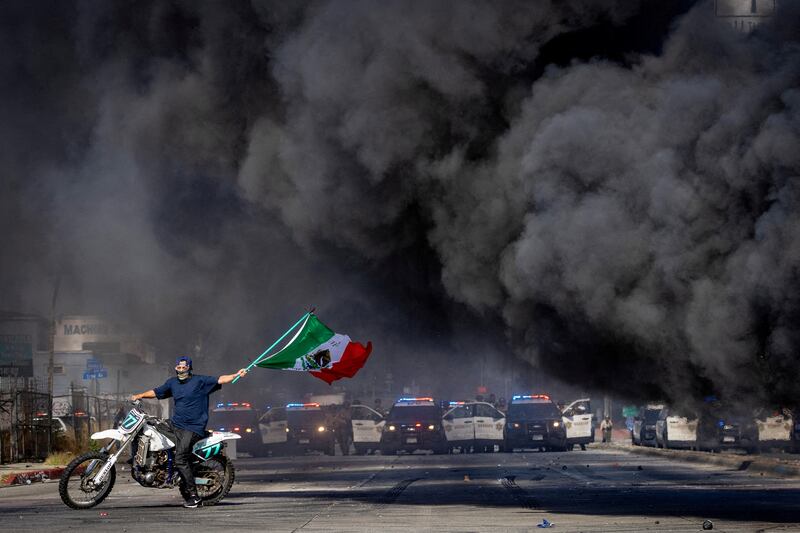 A man on a motorcycle waves a Mexican flag as smoke rises from a burning car on Atlantic Boulevard, during a standoff by protesters and law enforcement, following multiple detentions by Immigration and Customs Enforcement (ICE), in the Los Angeles County city of Compton, California, U.S., June 7, 2025.