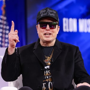 Elon Musk sits in a couch on stage the 2025 Conservative Political Action Conference wearing a dark hat, jacket and sunglasses.