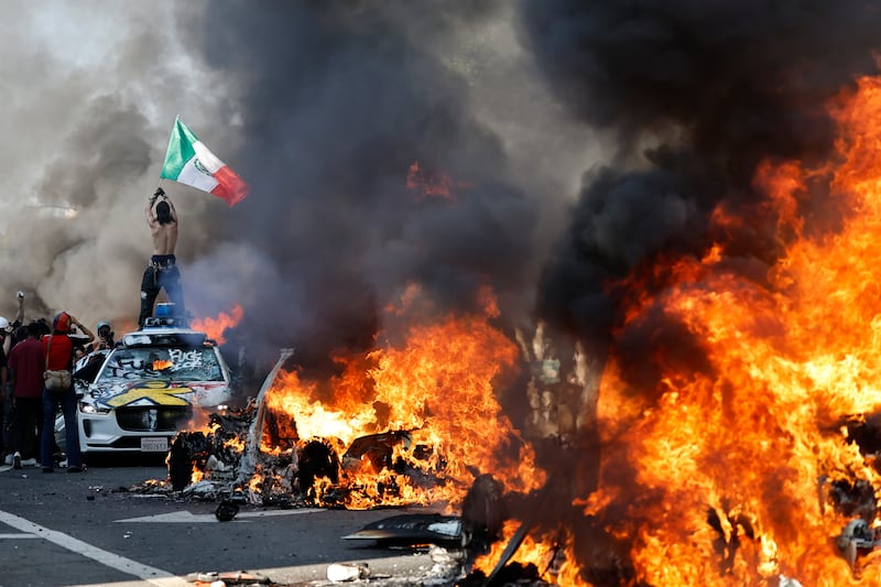 A protestor holds up a Mexican flag as burning cars line the street on June 08, 2025 in Los Angeles, California. Tensions in the city remain high after the Trump administration called in the National Guard against the wishes of city leaders following two days of clashes with police during a series of immigration raids.